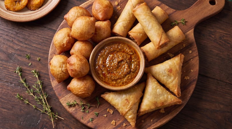 A wooden board with fried dough balls, spring rolls, samosas, and a bowl of spicy dip.