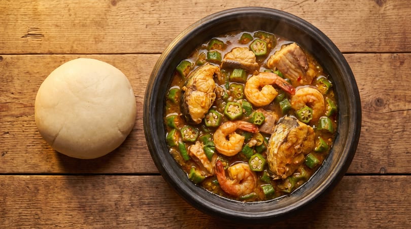 A bowl of okra soup with fish and shrimp, served with a side of fufu on a wooden table.