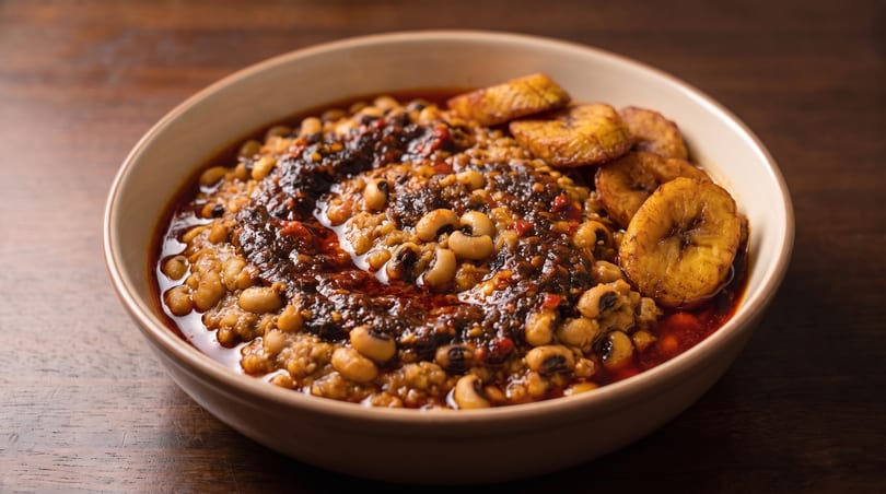 A bowl of black-eyed peas and stew, topped with fried plantains on a wooden table.