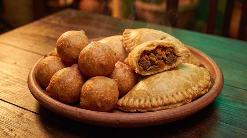 A plate of golden-brown fried dough balls and empanadas, one cut open to show a savory filling.